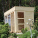 Traditional sauna cube with large windows situated in a garden surrounded by trees.