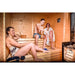 Traditional Sauna Cube 3 x 4m with Lounge with people relaxing inside and man using the ladle..