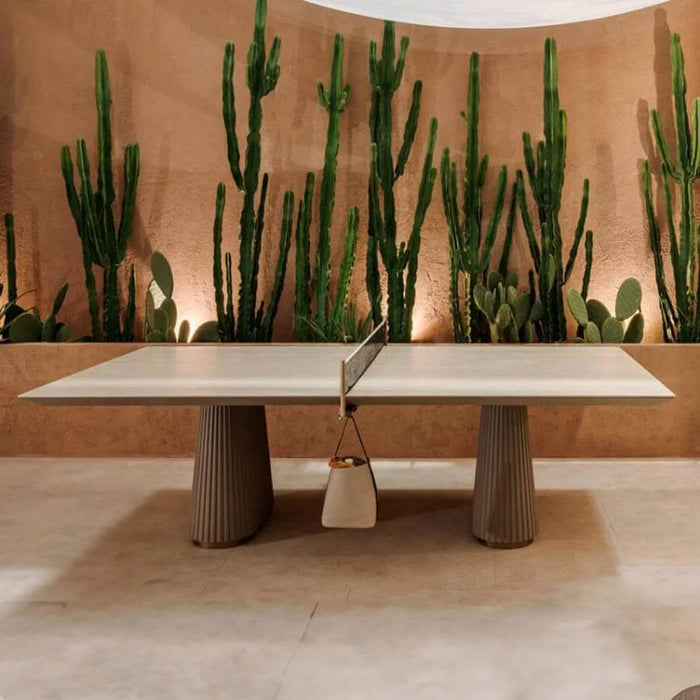 A front view of the Vismara Egeo Outdoor Table Tennis Table, featuring its Beige Travertino ceramic surface and fluted resin legs, elegantly placed against a warm backdrop of tall cacti.