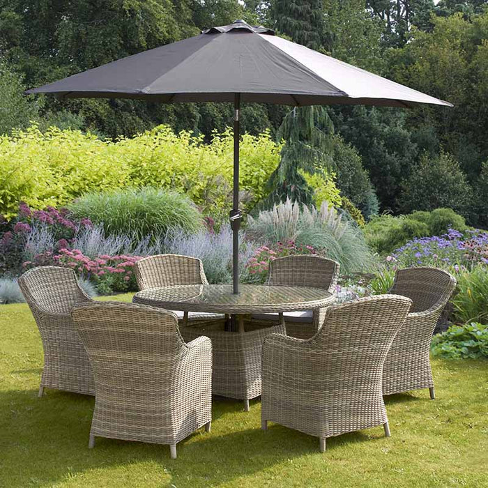 Wentworth 6 Seater Round Imperial Dining Set in a lush garden, showcasing a round glass-topped table and six rattan chairs under a large parasol.