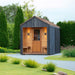 Woodnest Alice 2-3 Person Outdoor Sauna with covered timber entrance, glazed wooden door and dark cladding in a landscaped garden.