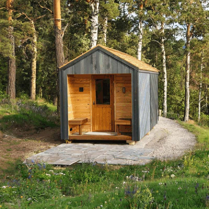 Woodnest Alice 2-3 Person Outdoor Sauna surrounded by woodland, featuring a natural timber porch, glazed door and grey wood exterior.