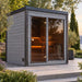 Woodnest Lucia 2 Person Outdoor Sauna with grey timber cladding, full-height glass front and warmly lit interior in a landscaped garden.