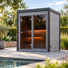 Woodnest Lucia 2 Person Outdoor Sauna beside a pool, with grey wood cladding, full-height glazing and warm interior lighting.