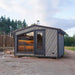 Woodnest Maria 2-4 Person Outdoor Sauna with grey timber cladding, large panoramic window and glass door in a woodland setting.